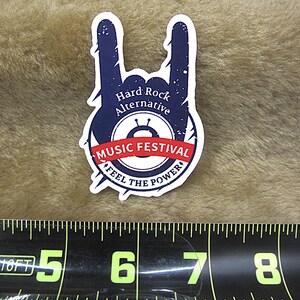 Puede incluir: Una pegatina azul marino con un diseño de gesto de mano de rock and roll. La pegatina tiene detalles en blanco y el texto "Hard Rock Alternative MUSIC FESTIVAL FEEL THE POWER". La pegatina mide aproximadamente 7,6 cm de ancho.