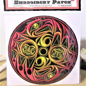 Può includere: Una toppa rotonda nera ricamata con un design rosso e oro. Il design presenta volti stilizzati e forme geometriche. La toppa ha il testo "EMBROIDERY PATCH" in alto e "MADE IN CANADA" in basso.
