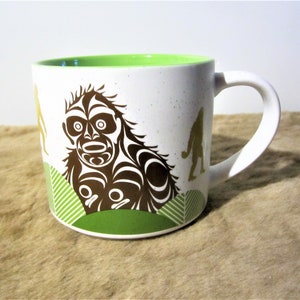 May include: White ceramic mug with a green rim and handle. The mug features a brown and white illustration of a stylised creature with a green background. The creature is surrounded by smaller, stylised figures in gold.