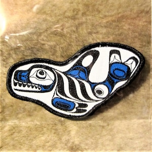 Può includere: Una toppa ricamata che raffigura una balena orca stilizzata in uno stile d'arte indigeno tradizionale della costa nord-occidentale, con colori bianco, nero e blu.