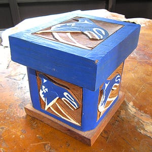 May include: A blue wooden box with carved whale designs. The box is square, painted blue, and sits on a natural wood base. The whale carvings are white and natural wood.