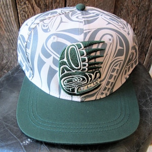 May include: A baseball cap with a white and grey pattern, a green peak, and a green emblem. The emblem features a stylized design. The cap has a flat peak and a button on top. The background is a wooden surface.