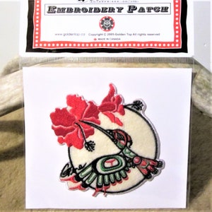 Può includere: Una toppa ricamata con un colibrì stilizzato in verde, rosso e nero, con un fiore rosso sullo sfondo. La toppa è su uno sfondo bianco e ha un bordo bianco. Il testo "EMBROIDERY PATCH" è stampato in nero nella parte superiore della toppa.
