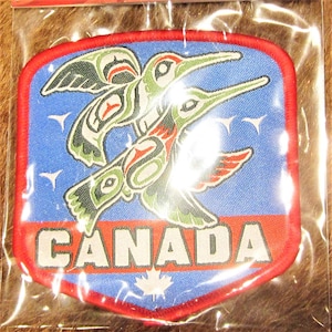 May include: An embroidered patch featuring two stylised birds in flight against a blue background. The patch is red, white and blue and has the word "CANADA" in white lettering and a maple leaf in the bottom corner.