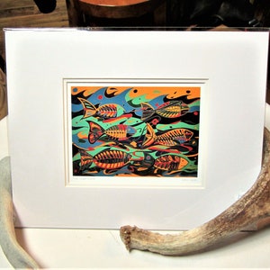 Op de afbeelding: Een ingelijste print van een kleurrijk abstract kunstwerk dat vissen toont die zwemmen in een turquoise en oranje oceaan. De vissen zijn gestileerd en hebben een skeletachtig uiterlijk. De print is getiteld "Fish Swim Swim".