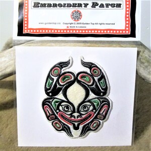 Può includere: Una toppa ricamata con una creatura stilizzata di colore nero, rosso e verde su sfondo bianco. La toppa è circondata da un bordo bianco e ha il testo "EMBROIDERY PATCH" nella parte superiore.