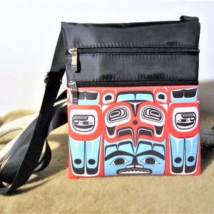 May include: Black crossbody bag with a red, blue and black patterned front flap. The flap features a stylised design of a face with geometric shapes.