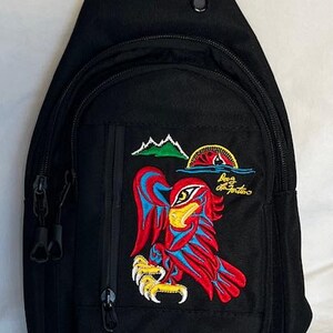 May include: Black canvas sling bag with an embroidered red, yellow and blue eagle design. The bag has a zipped pocket on the front and a zipped closure at the top. The text "Boca da Fortuna" is embroidered on the bag.