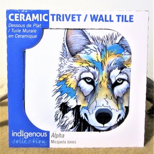 May include: Ceramic trivet or wall tile featuring a watercolor-style illustration of a wolf's face. The wolf has blue, yellow, and gray fur. The tile is part of the Indigenous Collection by CAP and is titled "Alpha" by Micqaela Jones.