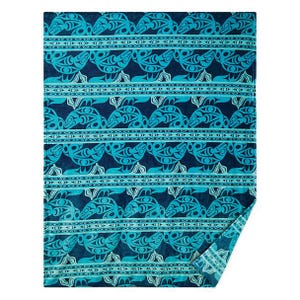 May include: A folded blanket featuring a repeating pattern of teal and navy blue turtle motifs. The blanket has a soft texture, with a folded corner displaying the pattern's edge.