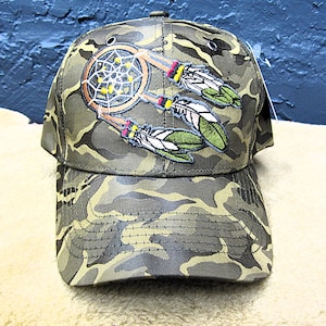 May include: Camouflage baseball cap with a dreamcatcher and feather design. The cap features a green, brown, and tan camouflage pattern. The dreamcatcher is embroidered with a tan circle, yellow web, and tan and red accents. The feathers are green and white.