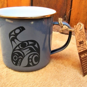 May include: A blue enamel mug featuring a black and white orca design. The mug has a gold rim and a black handle. The mug is tied with twine to a brown tag that reads "ORCA Maynard Johnny & Kwakwakwabkwakw ERDEL MUG This 12 oz mug is perfect for tea or coffee in your garden on a boat or camping. It is also a good size for a hearty mug of soup by the campfire."