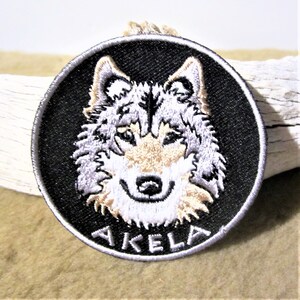 Peut inclure: Un écusson brodé noir et blanc représentant la tête d'un loup avec le mot "AKELA" en dessous. L'écusson est circulaire et a un fond noir.