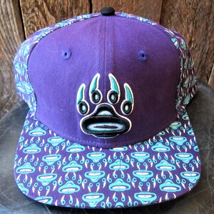 May include: Purple and teal baseball cap with a bear paw print on the front and a teal and white pattern on the brim.