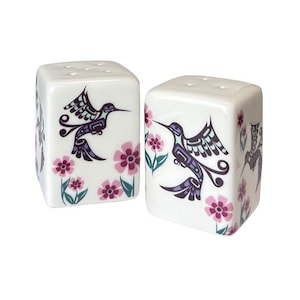 May include: A pair of white ceramic salt and pepper shakers. Each shaker features a stylized hummingbird design in purple and black, with pink flowers and green leaves. The shakers have a square shape with holes on top.