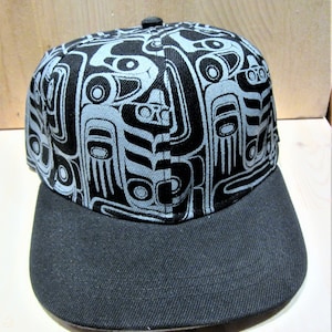 Könnte beinhalten: Eine schwarze Baseballkappe mit grauem Muster und schwarzem Schirm. Das Muster zeigt ein stilisiertes Design eines Vogels oder Tieres.