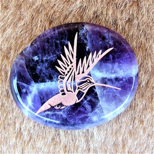 May include: A purple amethyst stone with a copper hummingbird etched into it. The stone is smooth and polished, and the hummingbird is detailed and intricate.