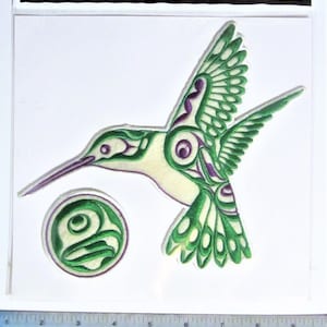 May include: Green and purple embroidered patch of a hummingbird with a stylized design. The patch is on a white background with a ruler for scale.