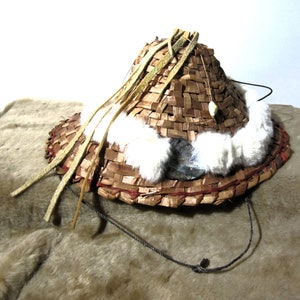 May include: A woven straw hat with a wide brim, trimmed with white fur and leather straps. The hat has a decorative piece of shell attached to the front.
