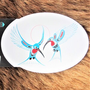 May include: A white oval sticker with two blue hummingbirds with red accents. The hummingbirds are facing each other with their wings spread out. The sticker has a white background with a light blue circle in the center.