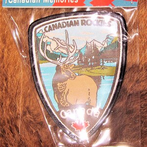 May include: A brown and black embroidered patch with a Canadian Rockies design. The patch features a large elk standing in a lake with mountains in the background. The text "CANADIAN ROCKIES" is printed in white on a black background. The text "CANADA" is printed in white on a brown background. A red maple leaf is printed in the bottom centre of the patch.