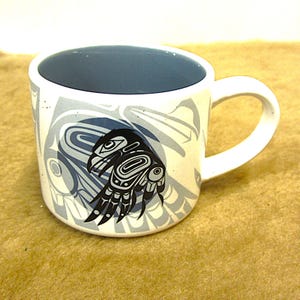 May include: White ceramic mug with a blue interior and a black and white stylized raven design. The raven is in a circle with a gray background. The mug has a white handle.