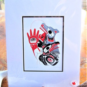 May include: A framed print of a Native American art piece depicting a person wearing a wolf mask and a raven headdress. The artwork is done in red, black, and grey colours.