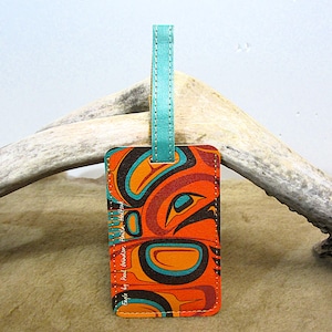 May include: A rectangular luggage tag with a teal leather strap. The tag displays a colourful orange, teal, and black design with the text "Eagle by Paul Windsor Haida". The tag is likely made of leather or a similar material.