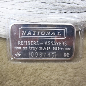 May include: A silver bar encased in clear plastic. The bar is rectangular with rounded corners and features the text "NATIONAL REFINERS - ASSAYERS" and "one oz troy SILVER 999+fine". A serial number and a maple leaf are also visible.