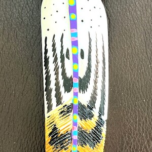 May include: A hand-painted feather with black, white, gold, purple, pink, blue, and yellow accents. The feather is painted with a geometric pattern and has a black tip.