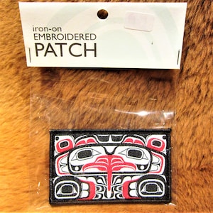 Könnte beinhalten: Aufbügel-Stickerei mit einem stilisierten Bärenkopf in Schwarz, Weiß und Rot. Das Patch ist in einer durchsichtigen Plastiktüte verpackt mit einem weißen Etikett, auf dem "iron-on embroidered patch" steht.