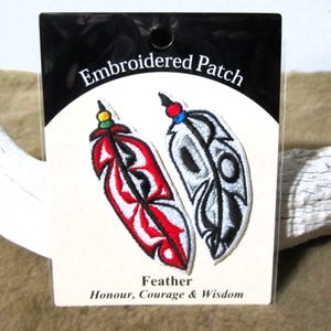 Può includere: Due toppe ricamate da stirare con piume. La toppa a sinistra è rossa, bianca e nera con una perlina verde e gialla in cima. La toppa a destra è grigia, bianca e nera con una perlina blu in cima. Le toppe sono confezionate su una carta bianca con il testo "Embroidered Patch" e "Feather Honour, Courage & Wisdom".