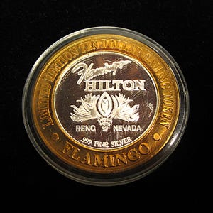 May include: A silver coin with a gold rim. The coin features the text "Limited Edition Ten Dollar Gaming Token" and "Flamingo Hilton" with a stylized flamingo design. The coin also says "Reno, Nevada" and "999 Fine Silver".