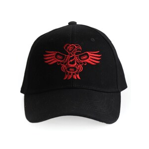 Può includere: Un berretto da baseball nero con un designo ricamato rosso di un uccello stilizzato. L'uccello ha un designo dettagliato con forme geometriche e linee.
