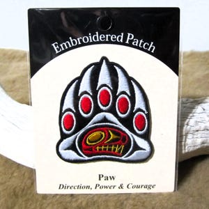 May include: Embroidered patch of a bear paw with red claws and a stylised bear face in the centre. The patch is on a white card with the text "Embroidered Patch" at the top and "Paw Direction, Power & Courage" at the bottom.