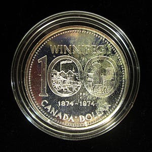 May include: A silver coin commemorating the 100th anniversary of Winnipeg, Canada. The coin features the text "Winnipeg 100 1874-1974 Canada Dollar" and depicts two scenes from the city's history.