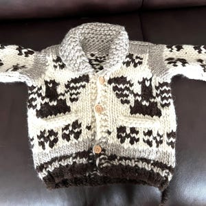 May include: A white and brown knitted cardigan with a button-up front and a contrasting brown collar. The cardigan has a geometric pattern with brown birds on a white background.