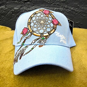 May include: Light blue baseball cap featuring a dreamcatcher design with pink roses and feathers. The cap has the words "Magic Pride" embroidered on the side. The cap is made of a light blue fabric.