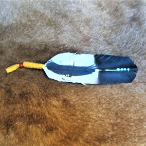 Peut inclure: Une plume d'aigle blanche et noire avec un motif d'aigle peint et des perles turquoise. La plume est attachée avec un cordon de cuir jaune et rouge.