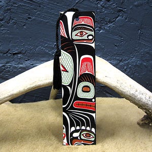 May include: A rectangular bookmark with a black tassel. The bookmark features a Native American-inspired design in black, red, white, and green. The artwork depicts stylized animal forms and geometric patterns. The bookmark is standing upright.