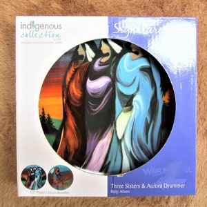 May include: A 7.5-inch ceramic plate featuring an Indigenous art design of three women in colorful clothing, with a sunset background. The plate is part of the Indigenous Collection by CAP and is titled "Three Sisters & Aurora Drummer" by Betty Albert.