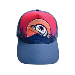 May include: A blue baseball cap with a curved brim. The cap features a design of a bird in a circle against a sunset backdrop. The design includes mountains and trees. The top of the cap is red and pink.