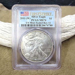 Peut inclure: Une pièce d'argent Silver Eagle de 2011 dans un étui de protection. La pièce présente un motif de Lady Liberty marchant et le texte "IN GOD WE TRUST". L'étui porte une étiquette avec "FIRSTSTRIKE" et "PCGS MS70".