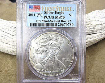 Könnte beinhalten: Eine silberne 2011 Silver Eagle Münze in einer Schutzhülle. Die Münze zeigt ein Design der wandernden Lady Liberty und den Text "IN GOD WE TRUST". Die Hülle hat ein Etikett mit "FIRSTSTRIKE" und "PCGS MS70".