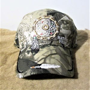 May include: A camouflage baseball cap with a white dreamcatcher and feather embroidery. The cap has the text "NATIVE PRIDE" embroidered on the side.