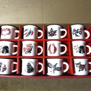 May include: A set of 12 white ceramic mugs with black and red designs. The mugs are arranged in a box with red and white lining. Each mug features a different design inspired by Indigenous art.