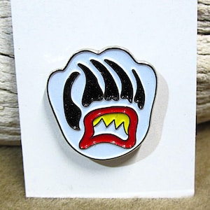 May include: A light blue enamel pin in the shape of a bear paw with black claws and a red and yellow mouth. The pin has a silver border and is mounted on a white card.