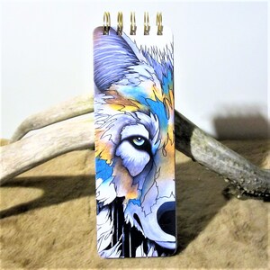 May include: A small spiral bound notebook with a colourful wolf illustration on the cover. The wolf's eye is blue and the fur is a mix of blue, yellow, and purple.