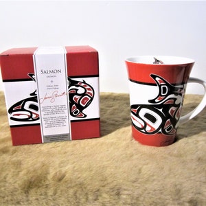 May include: A red and white ceramic mug with a black and red salmon design. The mug is in a box with a red background and a black and red salmon design. The box has the text "SALMON" and "Janice Stewart" on it.