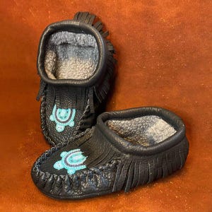 May include: A pair of black leather moccasins with turquoise beadwork depicting a turtle. The moccasins have fringe and are lined with grey fleece.